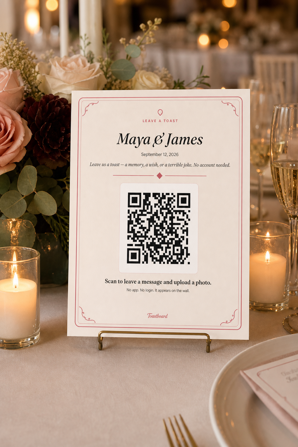 Printed cream Willow Book table sign with a rose border, Maya and James, and a large QR code standing on a guest table among candles and flowers